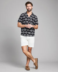 Man wearing a patterned black shirt, white shorts, and brown shoes on a gray background