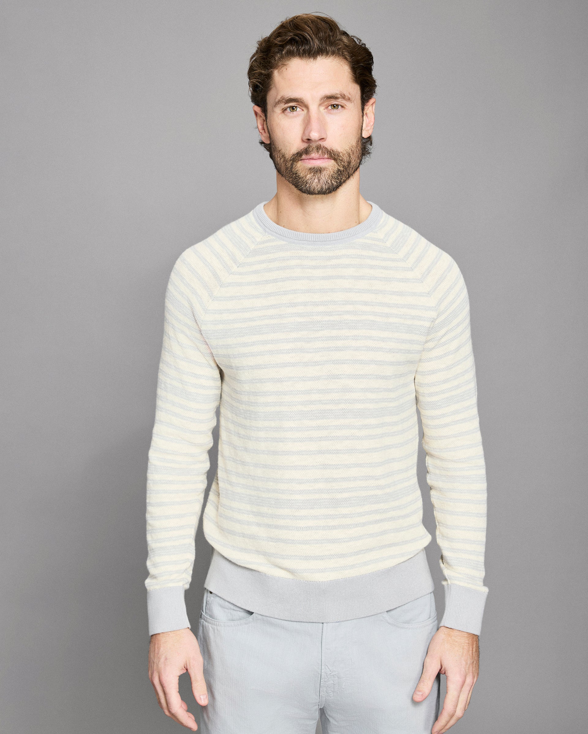 Man wearing a light-colored striped sweater and gray pants against a gray background