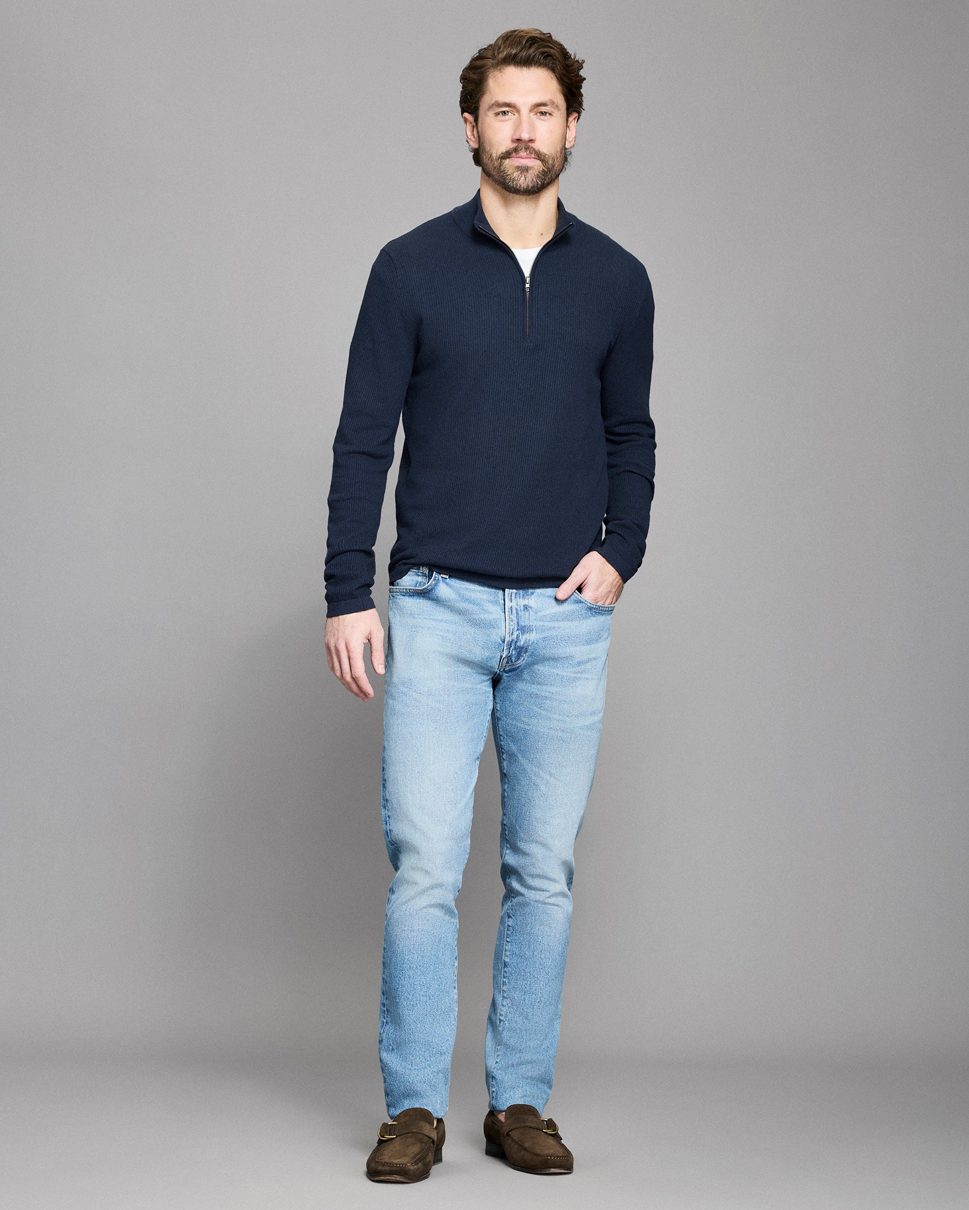 Man wearing a navy sweater and light blue jeans on a gray background