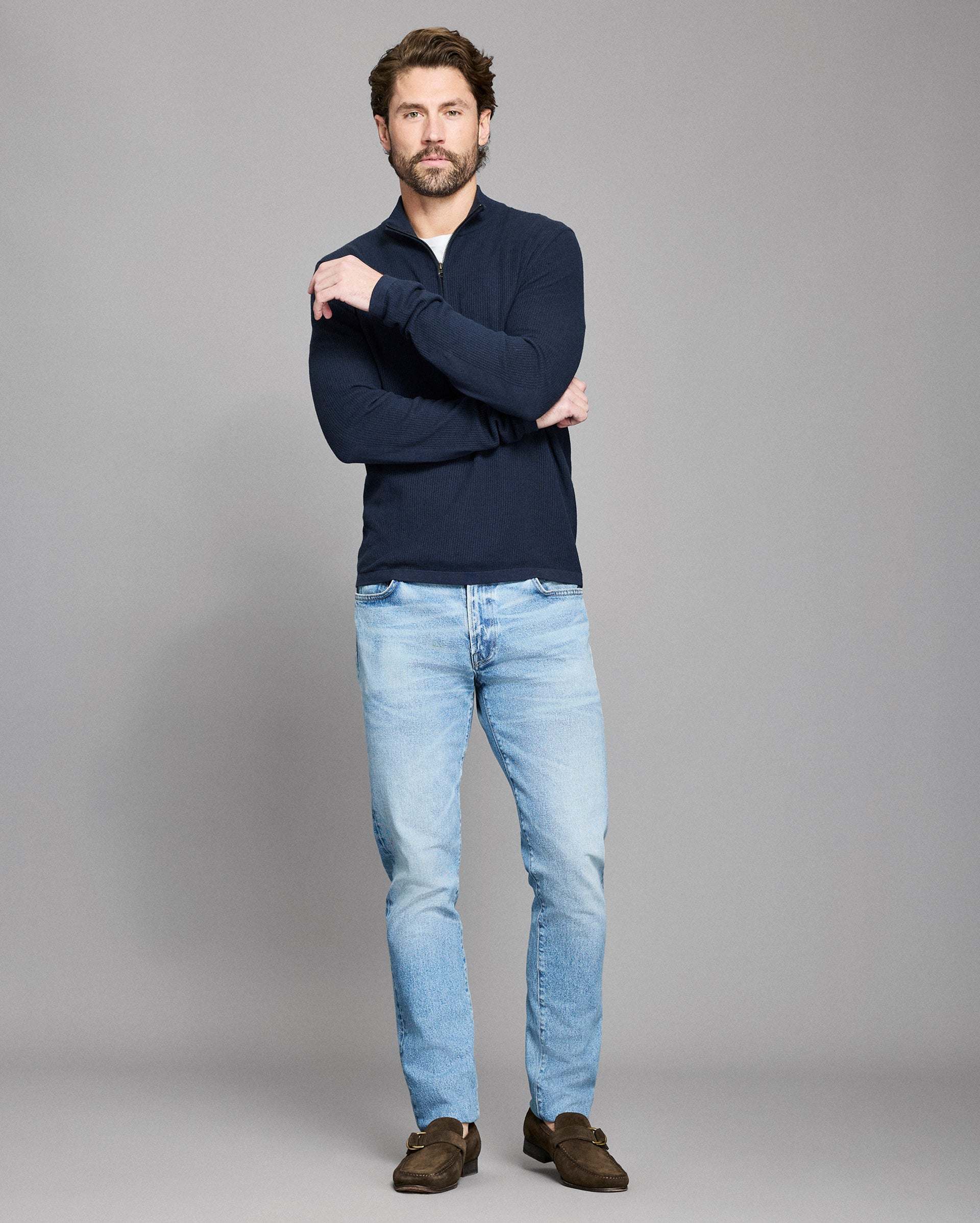 Man wearing a navy sweater and light blue jeans on a gray background
