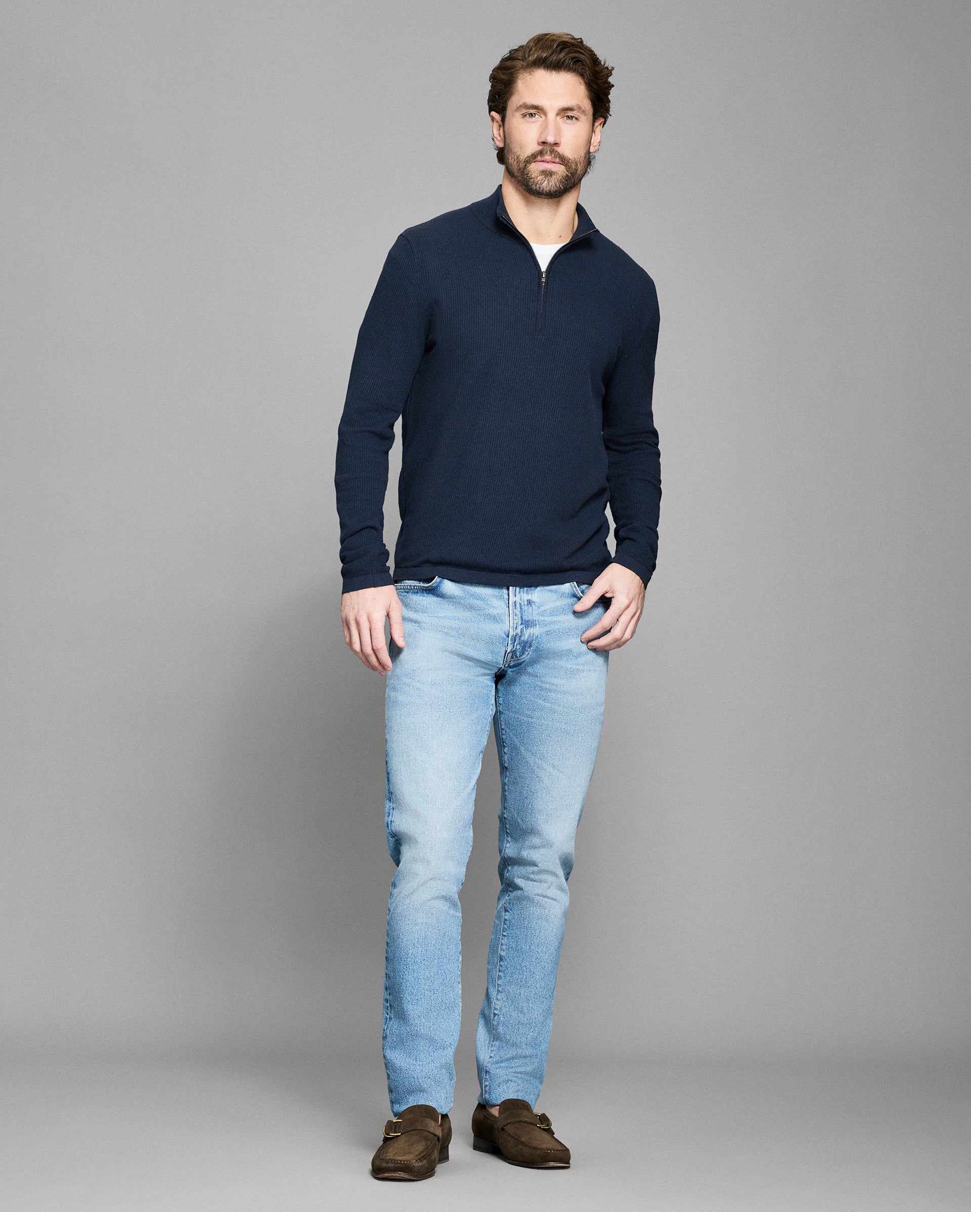Man wearing a navy sweater and light blue jeans against a gray background