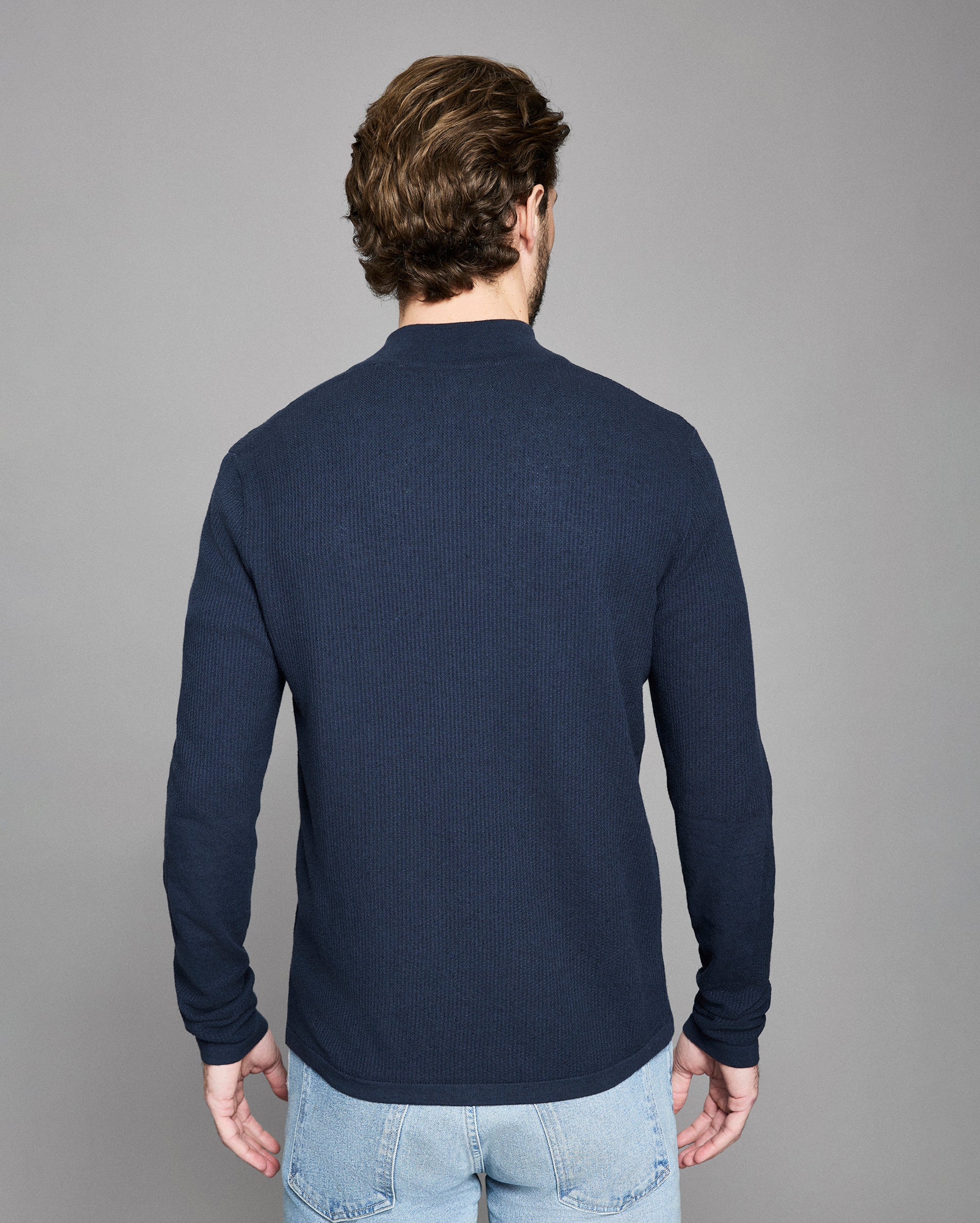 Man wearing a navy sweater and light blue jeans against a gray background