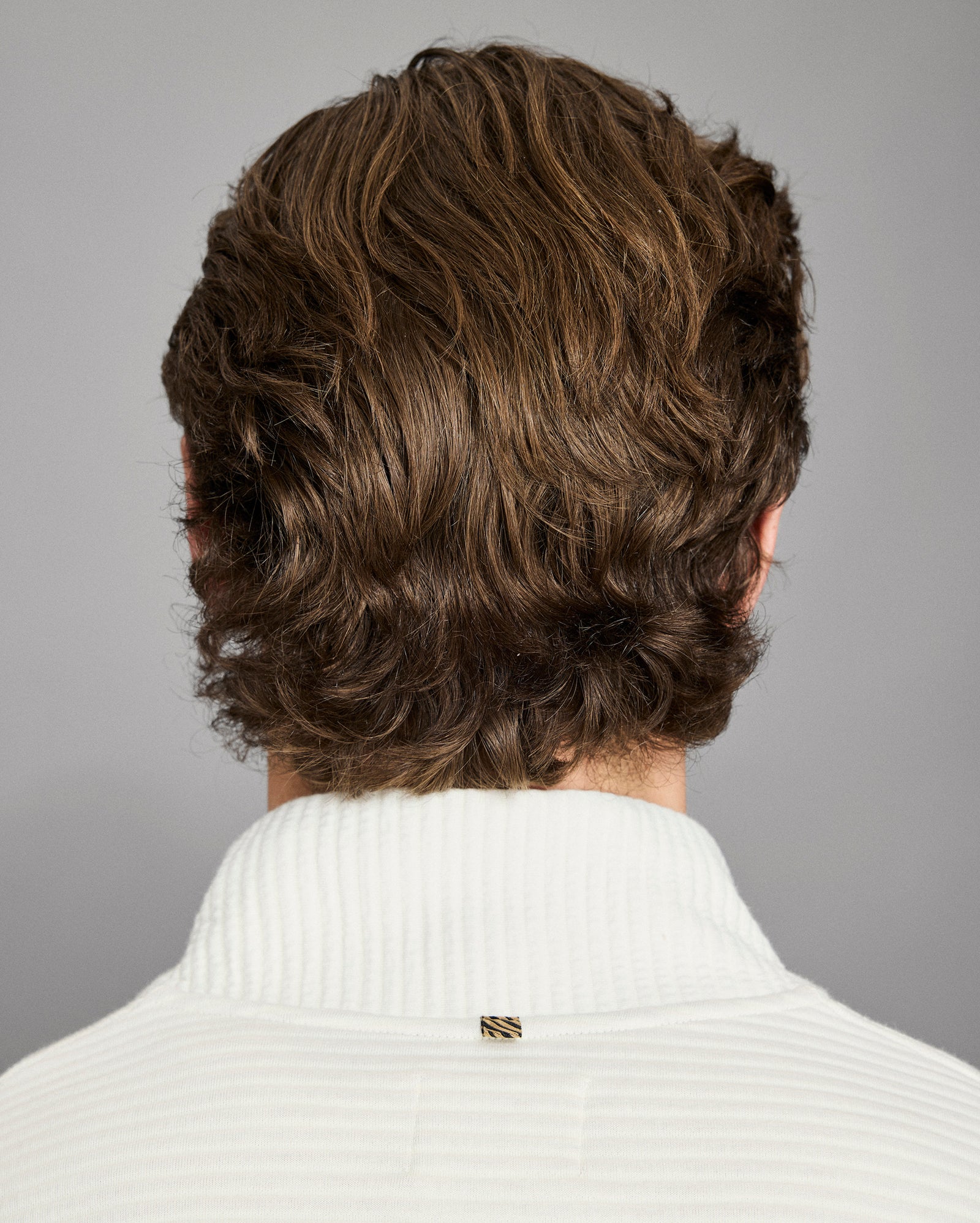 Back view of a person with wavy brown hair wearing a white sweater on a gray background