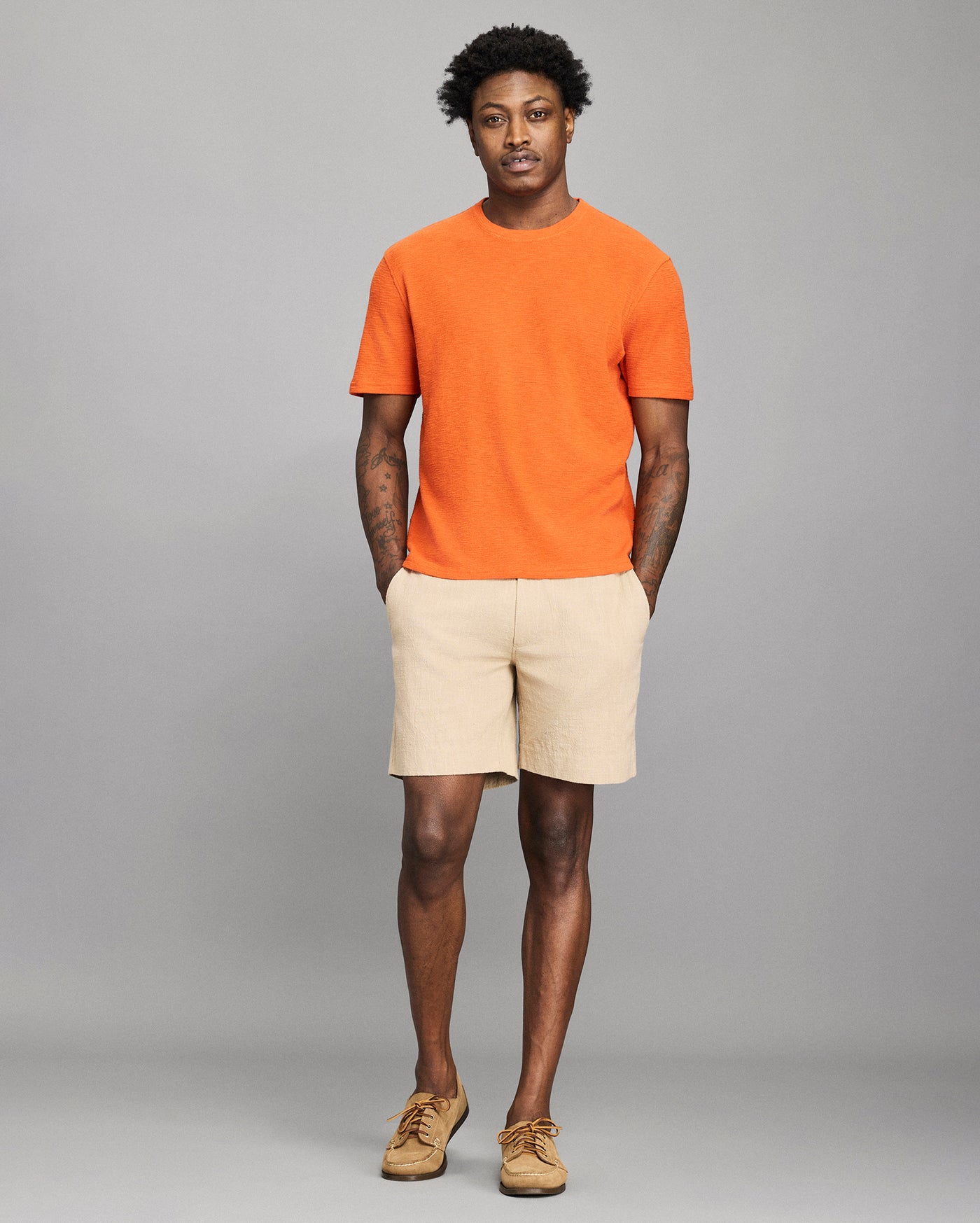Man wearing an orange t-shirt and beige shorts against a gray background