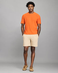 Man wearing an orange t-shirt and beige shorts against a gray background