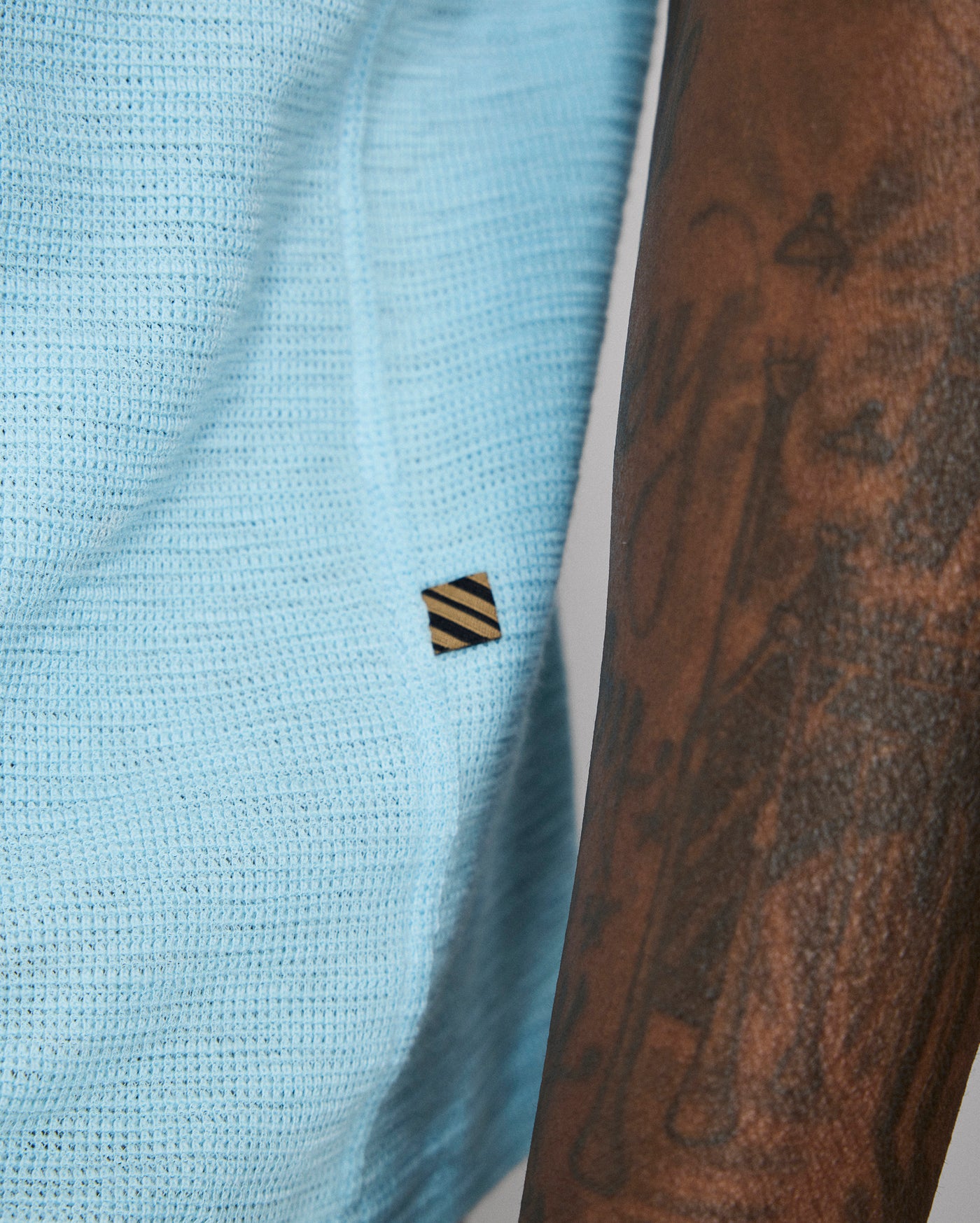 Light blue textured fabric with a small black and yellow striped logo, next to a tattooed arm.