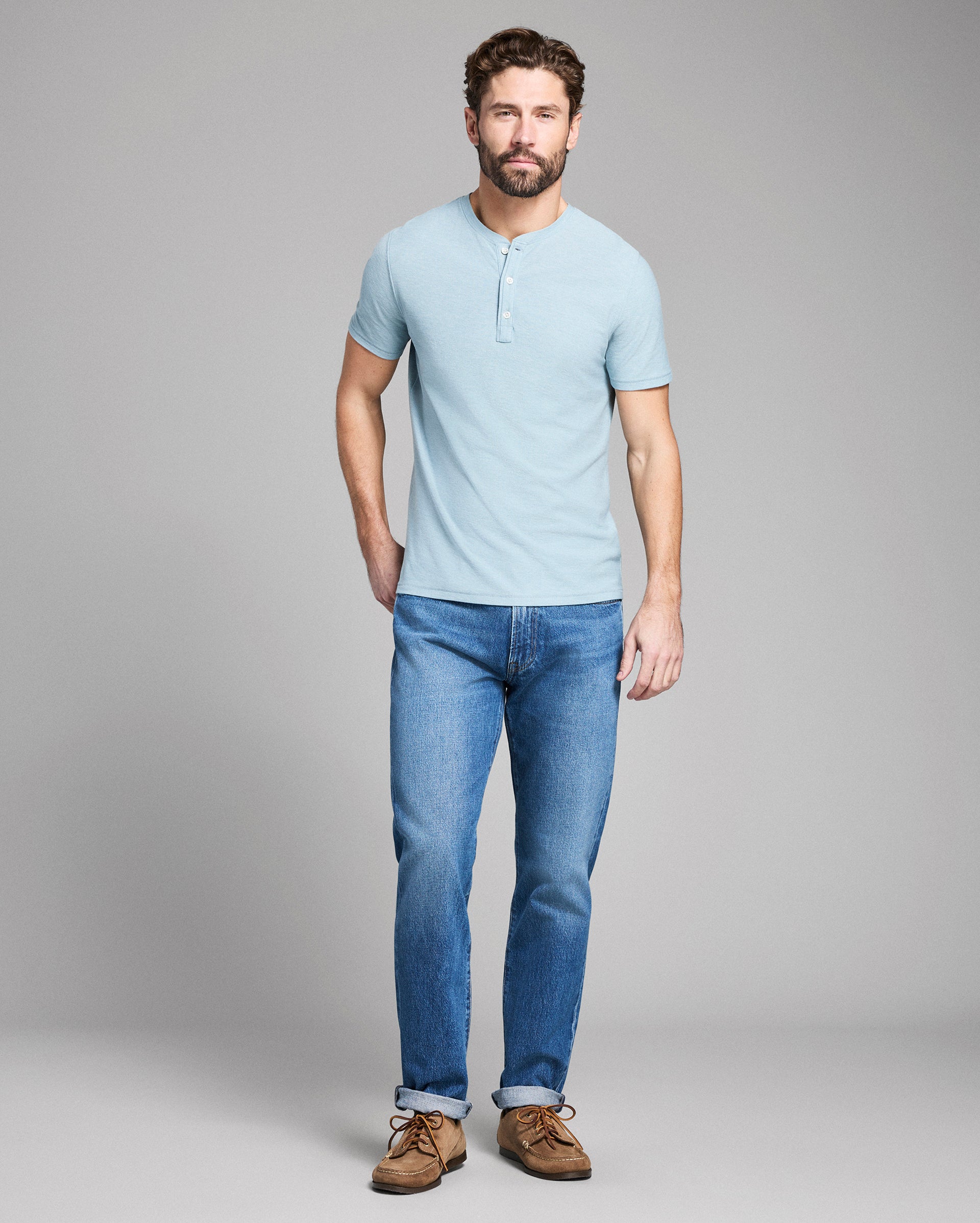 Man wearing a light blue henley shirt and jeans standing against a gray background