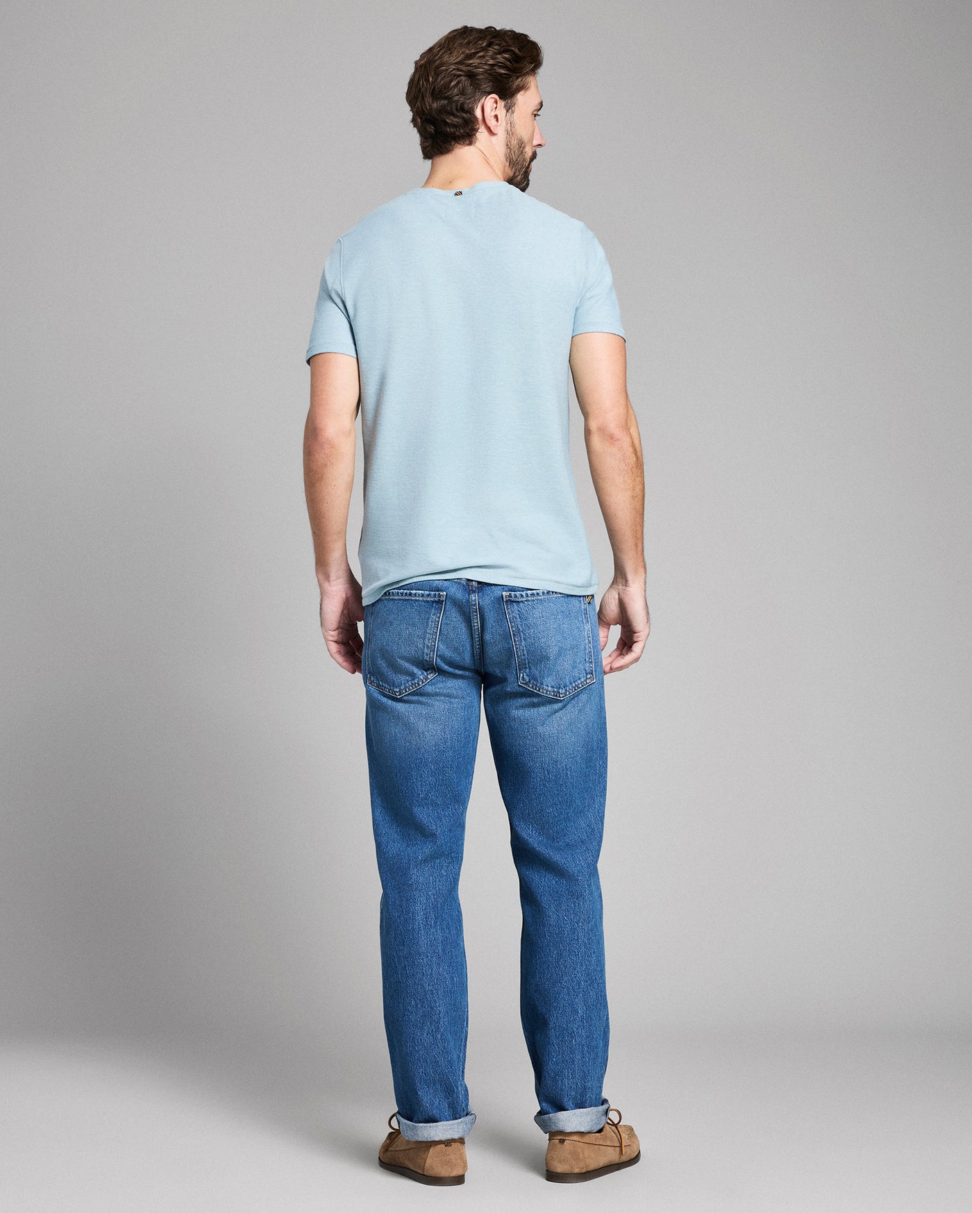 Man wearing a light blue t-shirt and blue jeans standing against a gray background