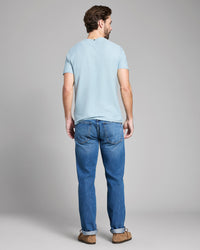 Man wearing a light blue t-shirt and blue jeans standing against a gray background