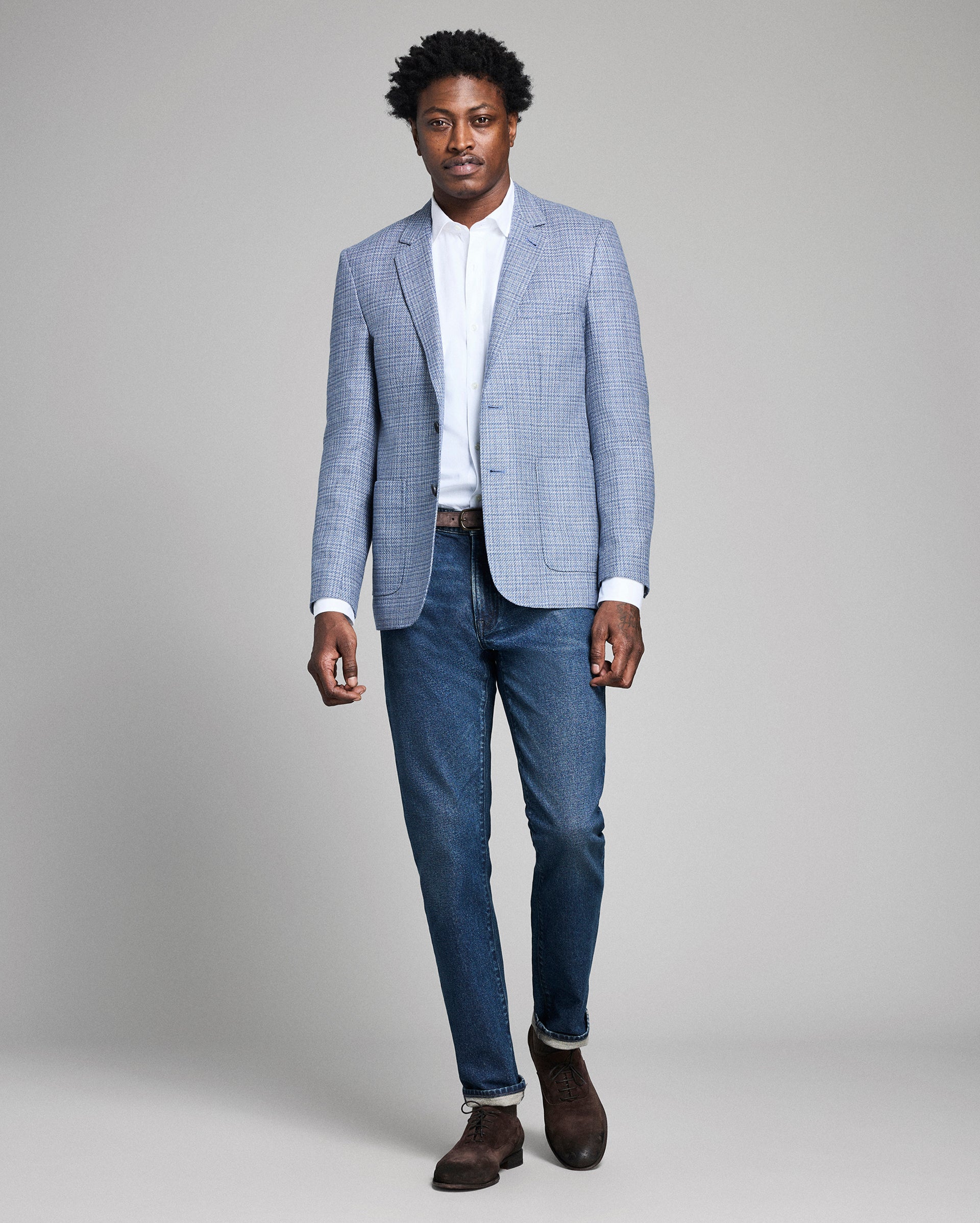 Man wearing a light blue checkered blazer, white shirt, and blue jeans on a gray background