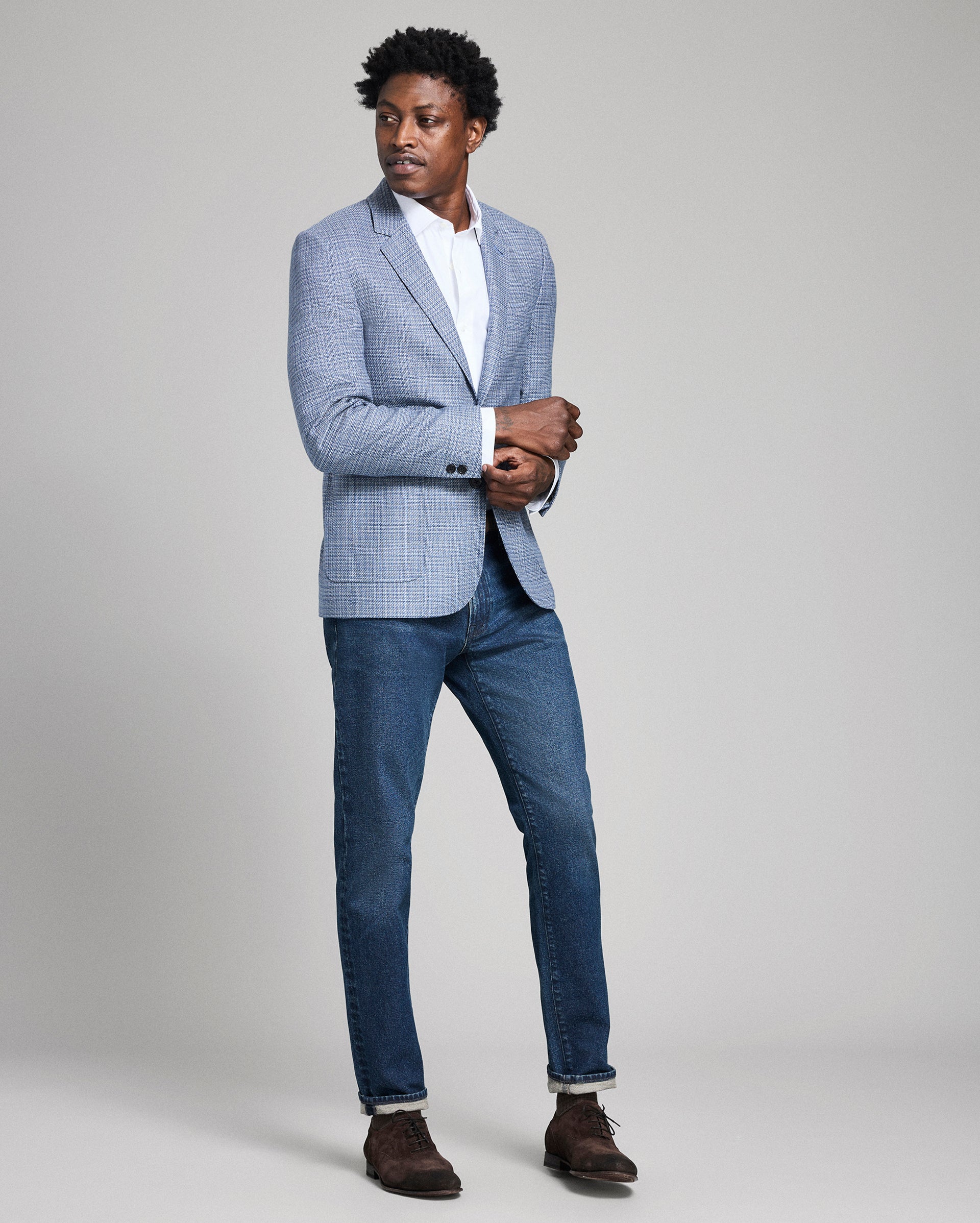 Man wearing a light blue checkered blazer, white shirt, and dark blue jeans on a gray background