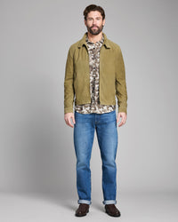 Man wearing a green jacket, camouflage shirt, and blue jeans on a gray background