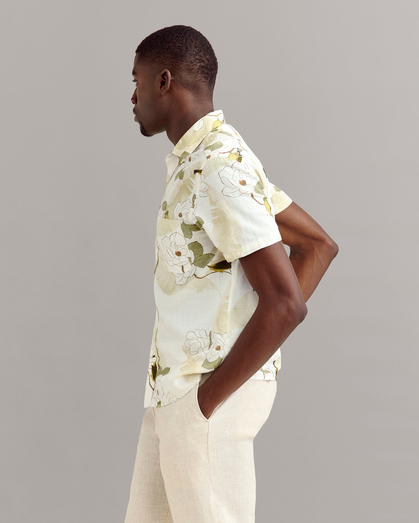 A man stands in profile against a plain gray background, wearing a light-colored, short-sleeve, floral button-up shirt and light beige pants, with his hands in his pockets.