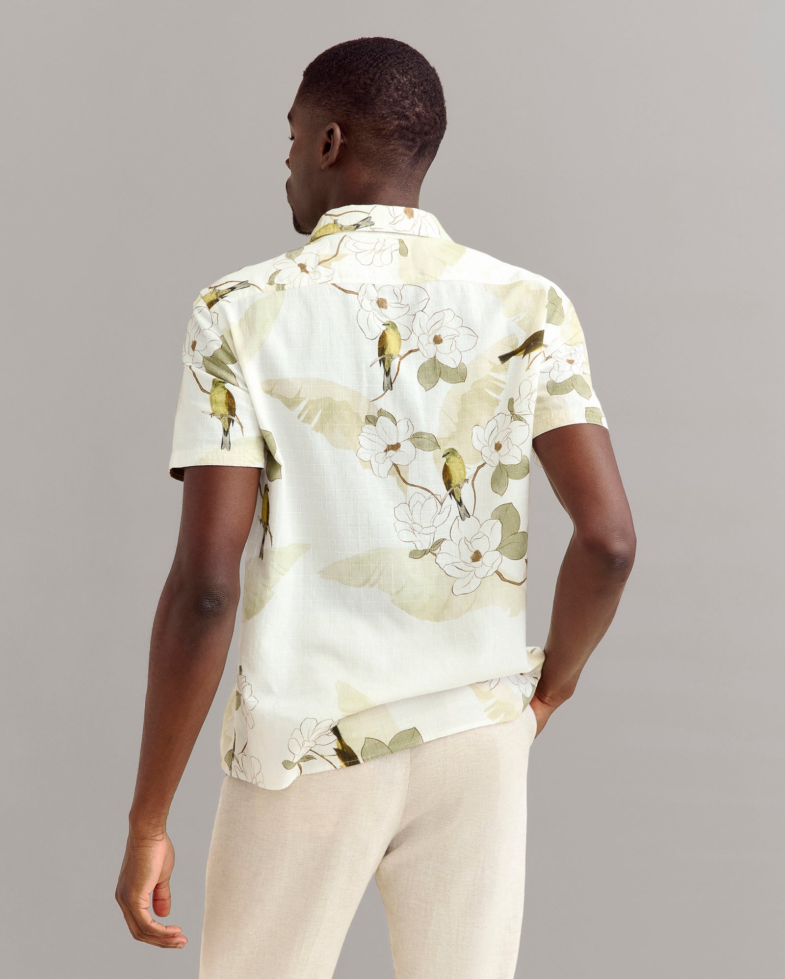 A man stands facing away, wearing a light-colored short-sleeve shirt with a floral and bird print, and beige pants against a plain, neutral background.
