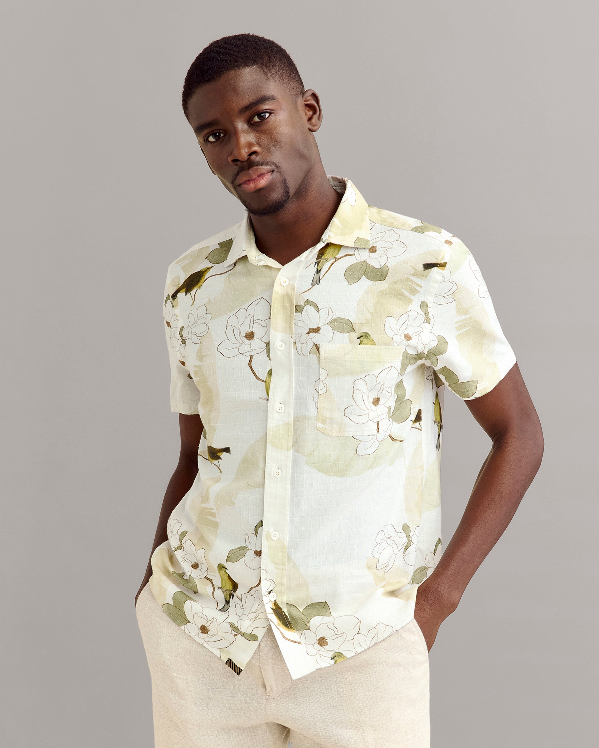 A man wearing a light-colored, short-sleeve button-up shirt with a floral pattern stands with his hands in his pockets against a plain gray background.