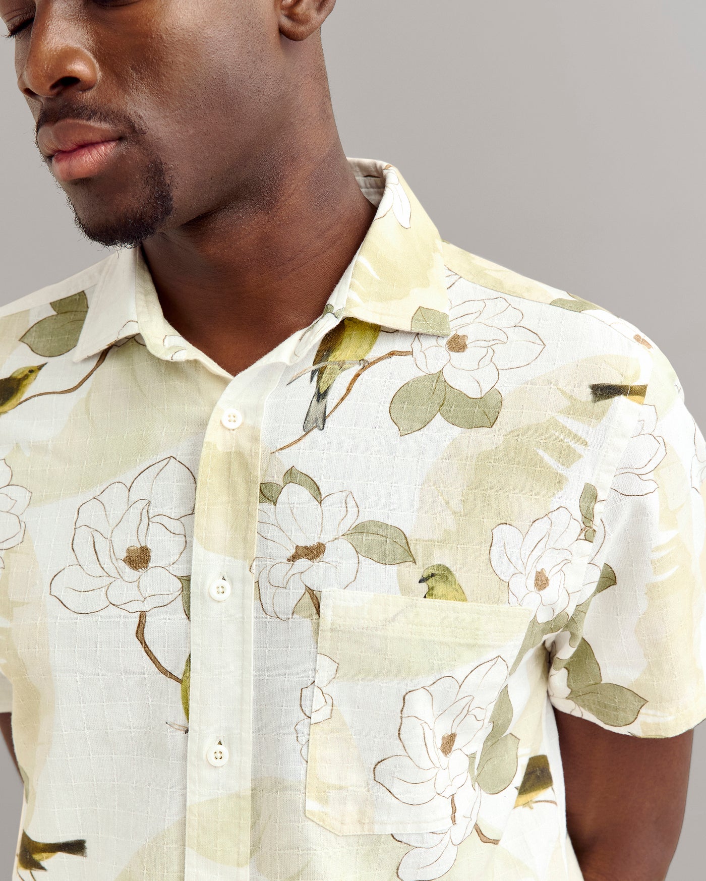 A man wearing a short-sleeved, light-colored button-up shirt with a floral pattern stands against a plain gray background. The image shows his face partially and highlights the shirts design.