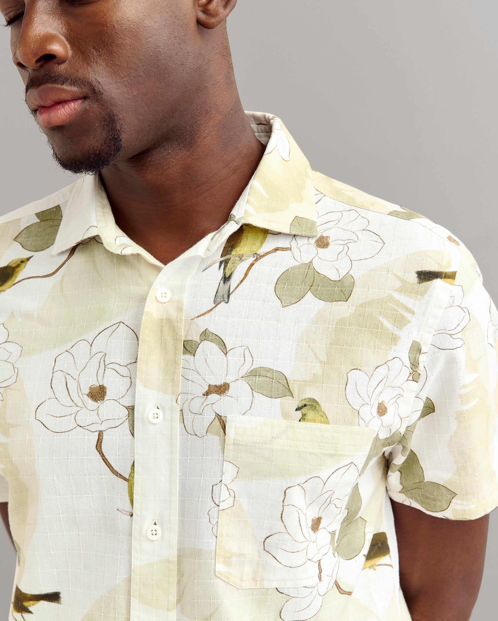 A man wearing a short-sleeved, light-colored button-up shirt with a floral pattern stands against a plain gray background. The image shows his face partially and highlights the shirts design.