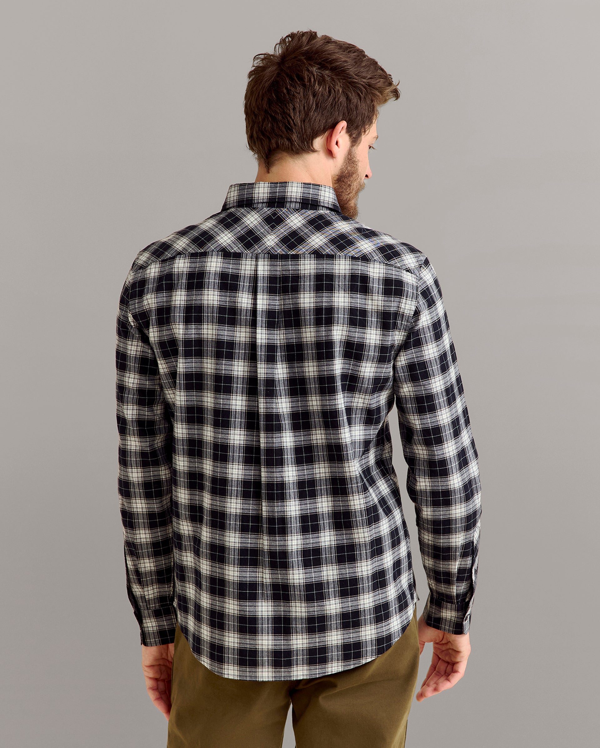 A man stands facing away from the camera, wearing a long-sleeve, black-and-white plaid shirt and olive-green pants against a plain light-gray background.
