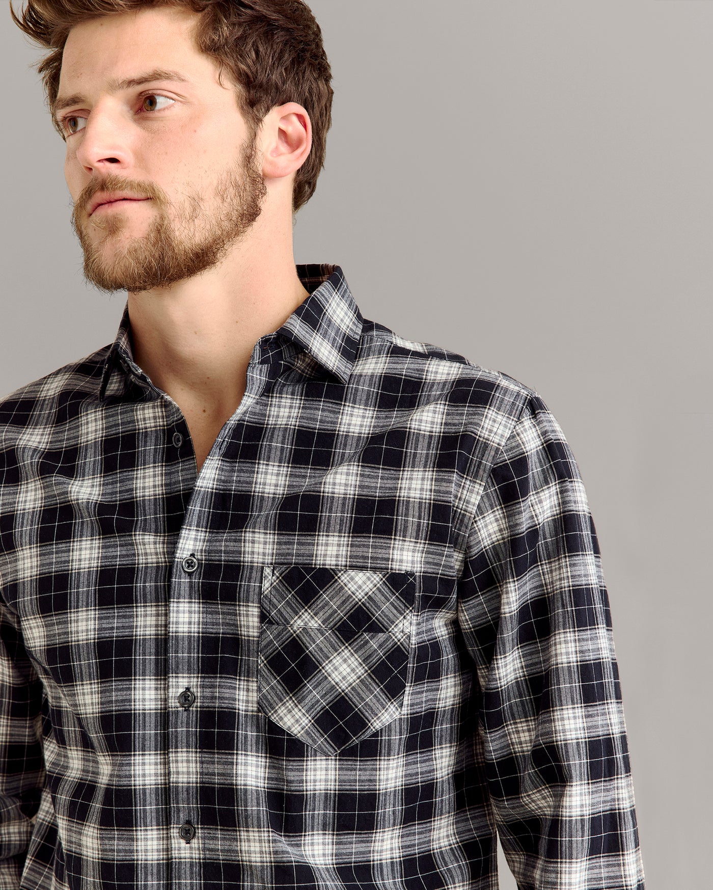 A man with short brown hair and a beard wears a black, white, and gray plaid button-up shirt with a front pocket, standing against a plain light gray background.