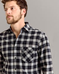 A man with short brown hair and a beard wears a black, white, and gray plaid button-up shirt with a front pocket, standing against a plain light gray background.