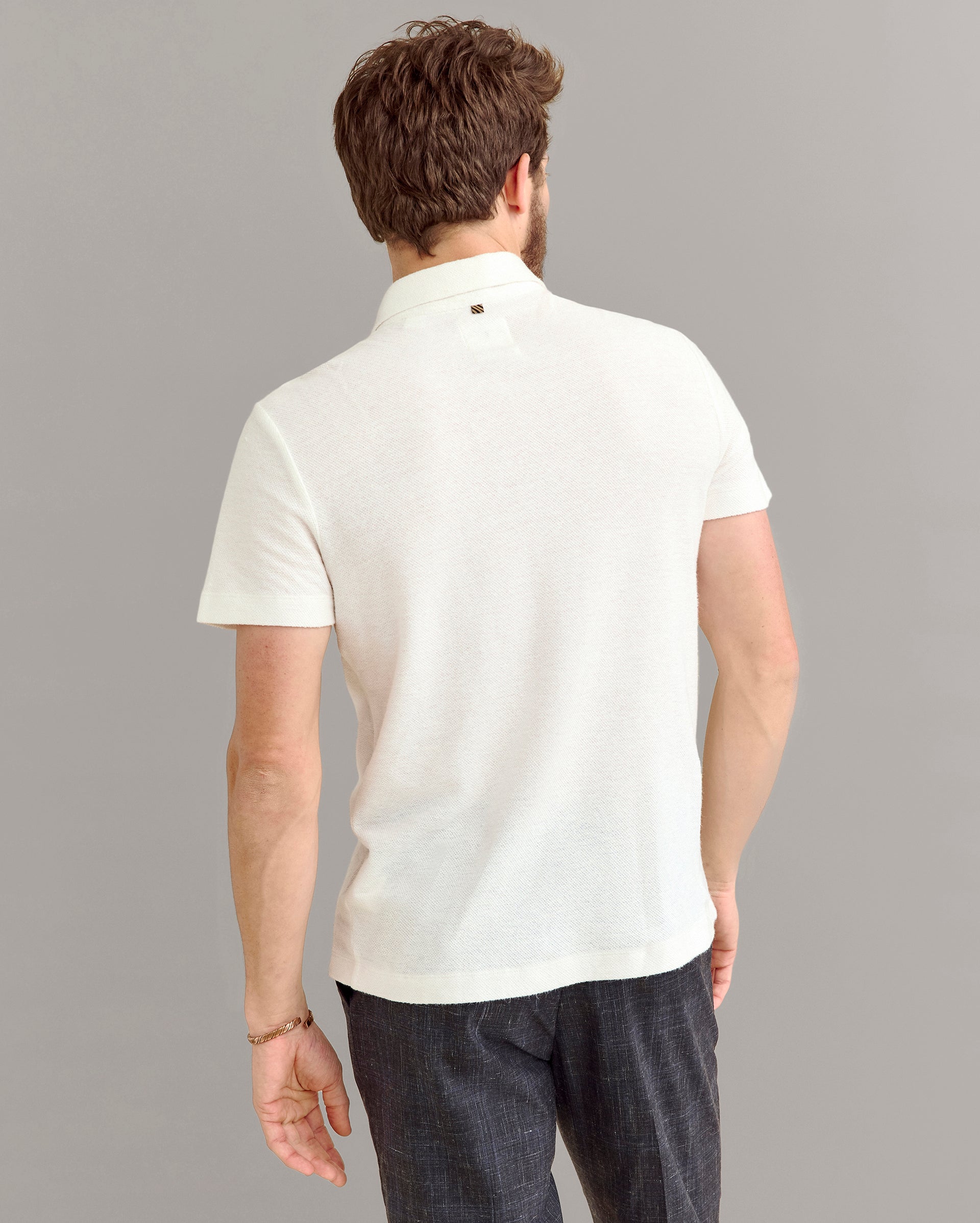 A man with brown hair is shown from the back, wearing a short-sleeved, off-white polo shirt and dark pants against a plain light gray background.