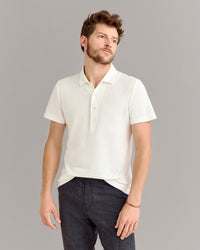 A man with short brown hair and a beard stands against a plain gray background, wearing a white short-sleeve polo shirt and dark pants, with one hand by his side and the other slightly bent.