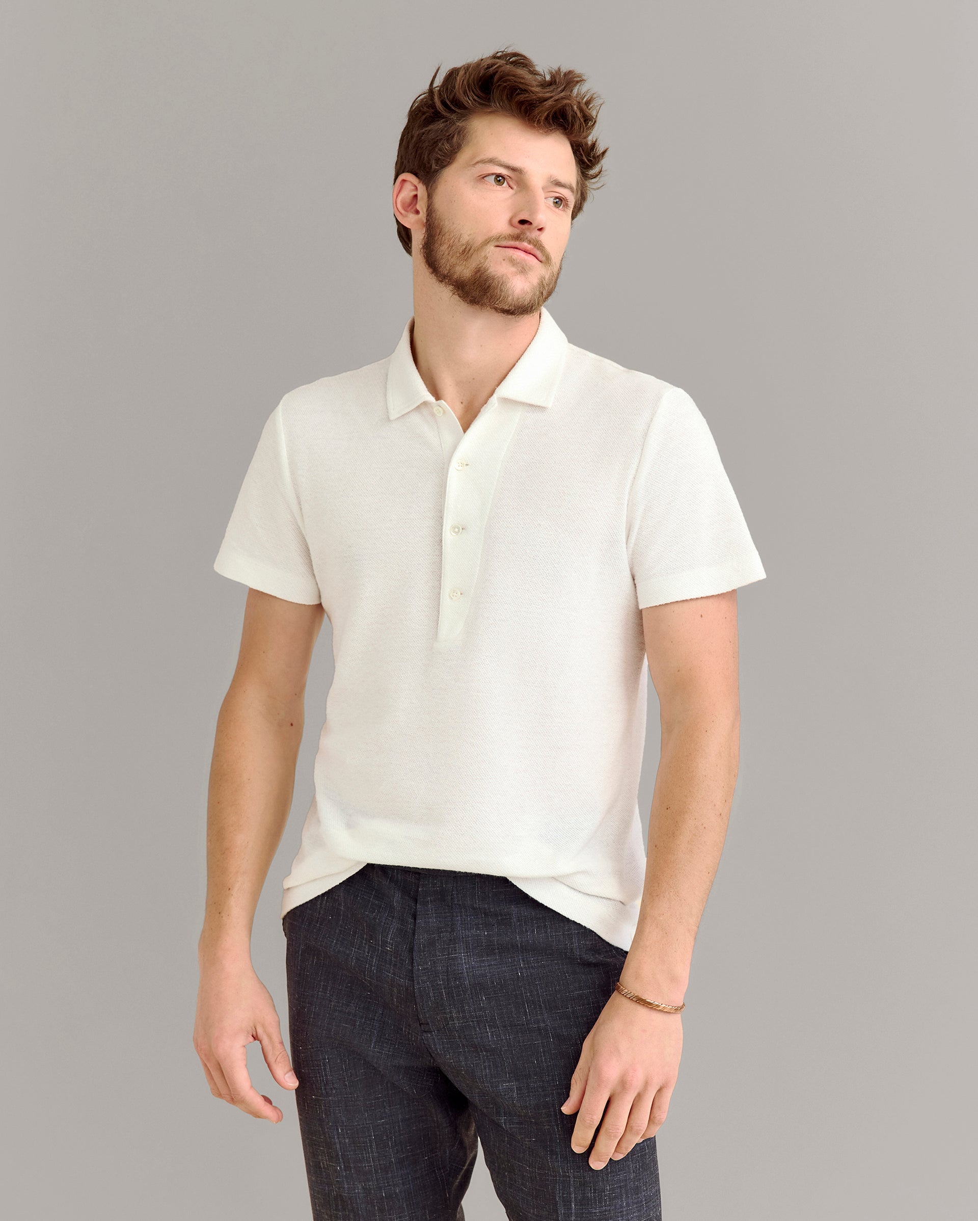 A man with short brown hair and a beard stands against a plain gray background, wearing a white short-sleeve polo shirt and dark pants, with one hand by his side and the other slightly bent.