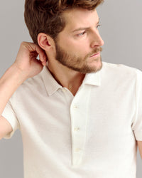 A man with short brown hair and a beard wearing a cream-colored short-sleeve polo shirt looks to his right, with one hand touching the back of his neck against a plain gray background.