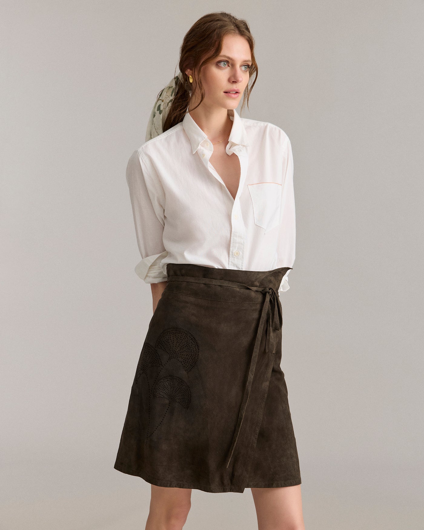 A woman wearing a white button-up shirt with rolled sleeves and a dark brown wrap skirt stands against a plain, light background, looking to the side with a neutral expression.