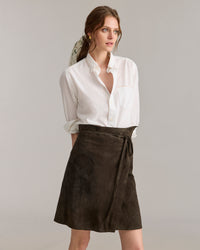 A woman wearing a white button-up shirt with rolled sleeves and a dark brown wrap skirt stands against a plain, light background, looking to the side with a neutral expression.