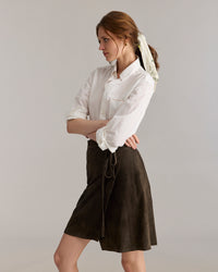 A woman with a light scarf in her hair wears a white button-up shirt with rolled sleeves and a dark wrap skirt, standing sideways with her arms folded against a plain background.