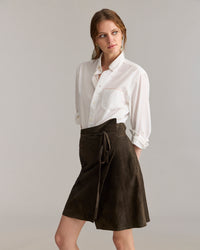 A woman stands against a neutral background, wearing a white button-up shirt with rolled sleeves and a dark brown wrap skirt tied at the waist. She looks slightly to the side with a relaxed expression.