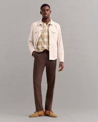 A man stands against a plain gray background wearing a cream jacket over a patterned beige shirt, dark brown pants, and tan suede shoes, with one hand in his pocket.