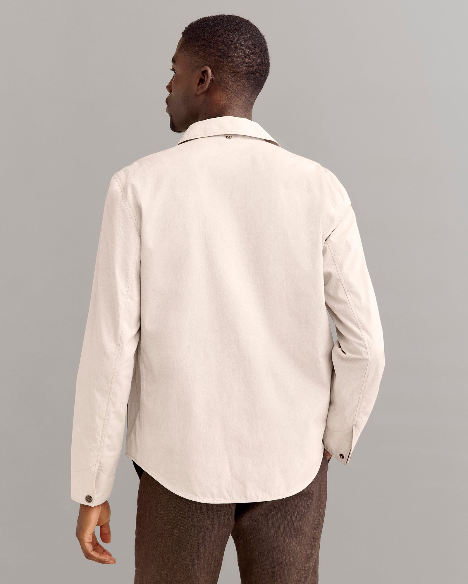 A man stands facing away from the camera, wearing a light beige long-sleeve button-up shirt and brown pants, against a plain gray background.