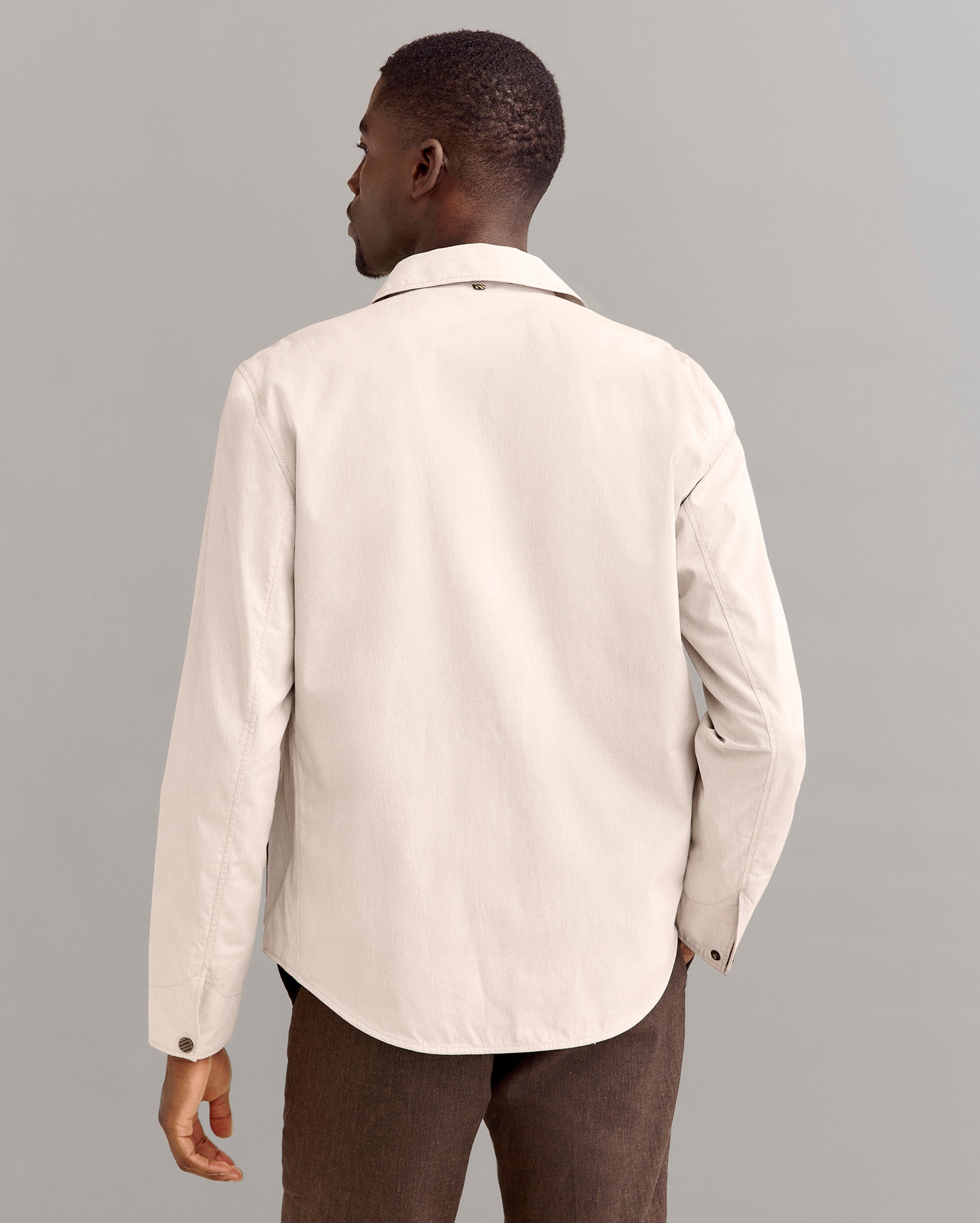 A man stands facing away from the camera, wearing a light beige long-sleeve button-up shirt and brown pants, against a plain gray background.