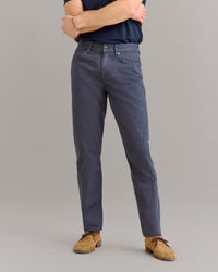 A person stands with arms crossed, wearing a navy T-shirt, gray slim-fit pants, and tan suede shoes, against a plain gray background. Their head is not visible.
