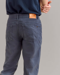 A person wearing blue pants and a dark blue shirt stands with one arm behind their back. The focus is on the back of the pants, which have patch pockets and a tan leather label on the waistband.