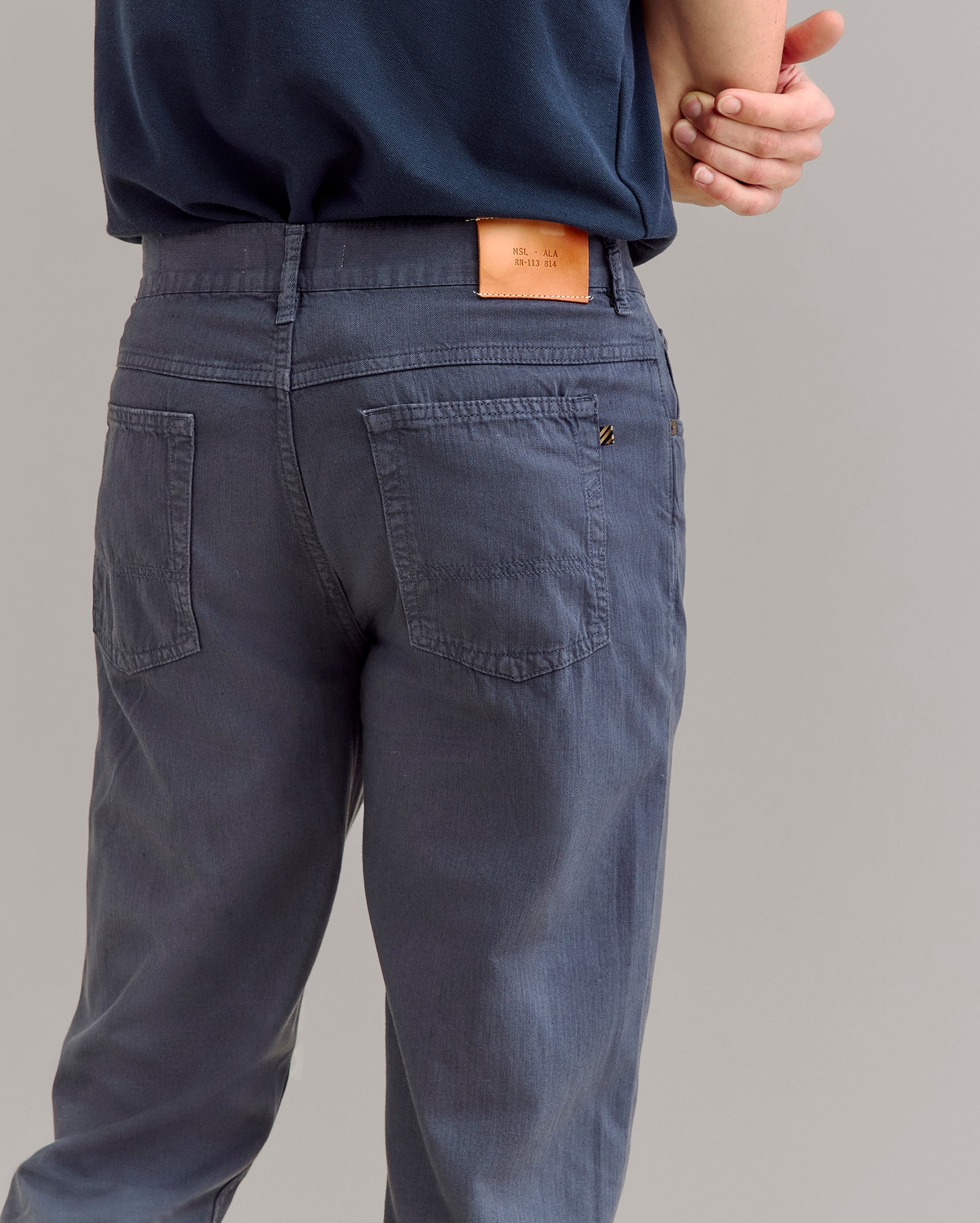 A person wearing blue pants and a dark blue shirt stands with one arm behind their back. The focus is on the back of the pants, which have patch pockets and a tan leather label on the waistband.