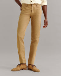 A person wearing light brown pants, a cream-colored shirt, and tan suede shoes stands against a plain gray background. Only the lower half of the body is visible.