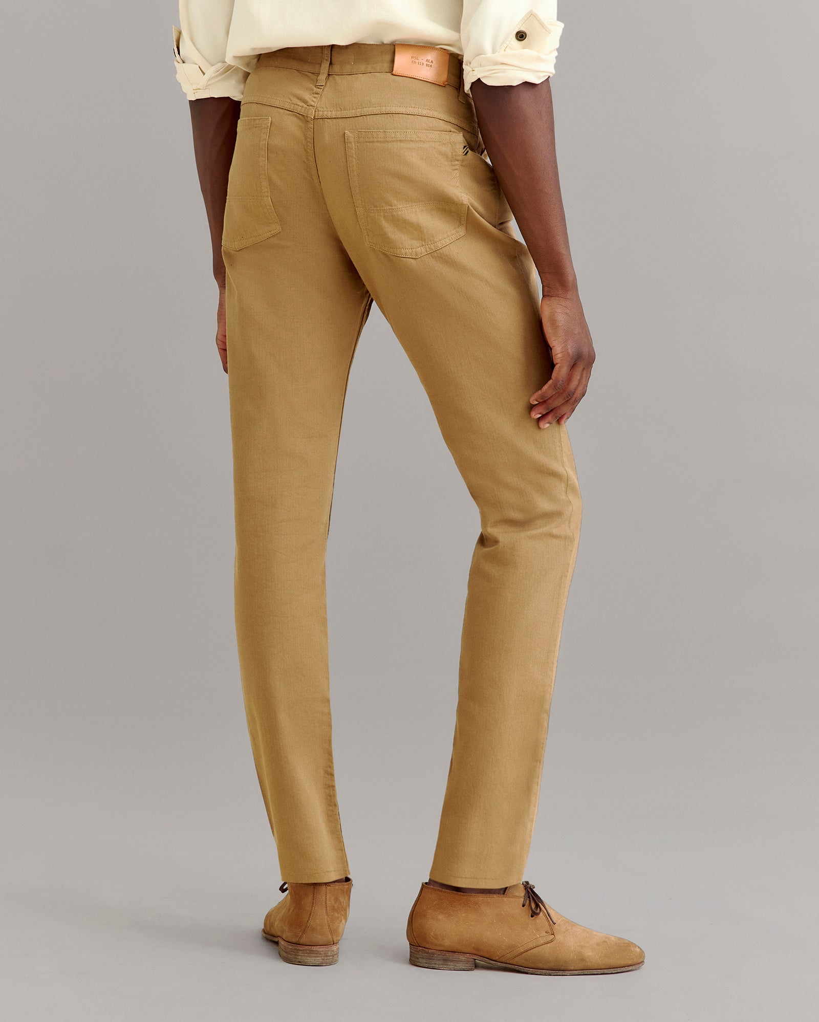 A person wearing tan slim-fit pants, a light-colored rolled-sleeve shirt, and brown suede shoes stands facing away, showing the back details of the pants against a plain gray background.
