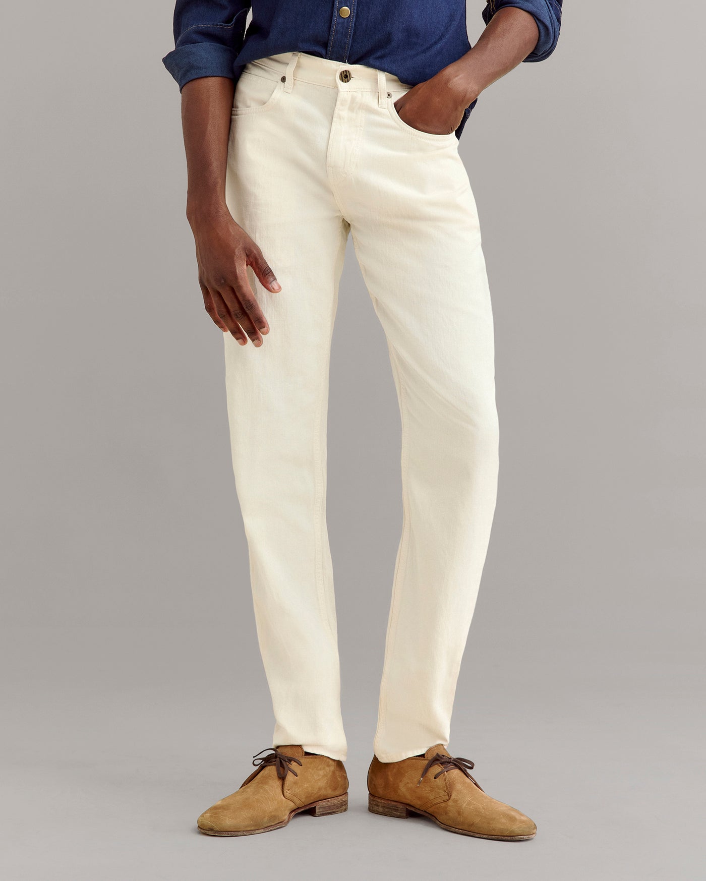 A person wearing cream-colored jeans, a dark blue button-up shirt, and tan suede shoes stands against a plain gray background, with one hand in their pocket.