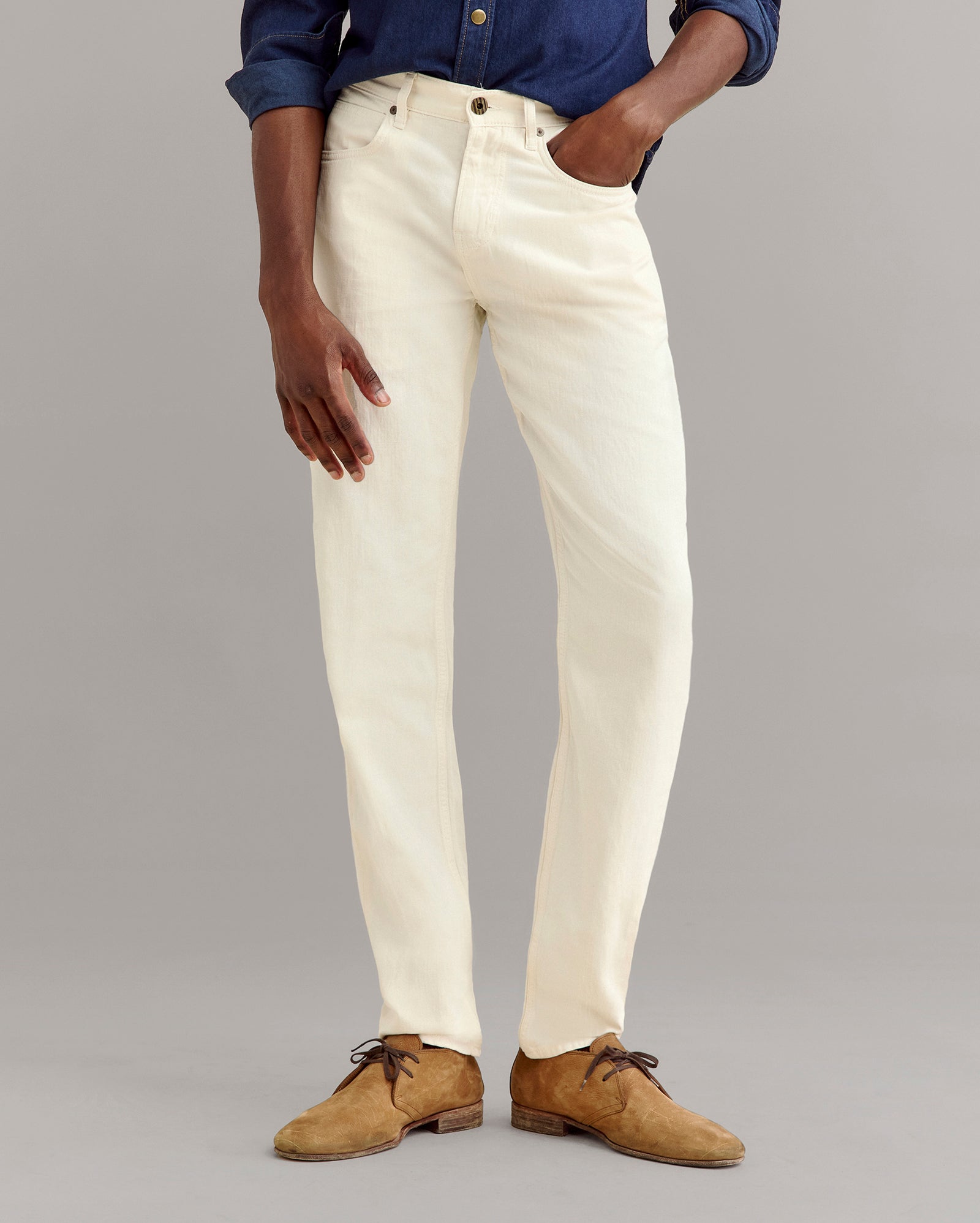 A person wearing cream-colored jeans, a dark blue button-up shirt, and tan suede shoes stands against a plain gray background, with one hand in their pocket.