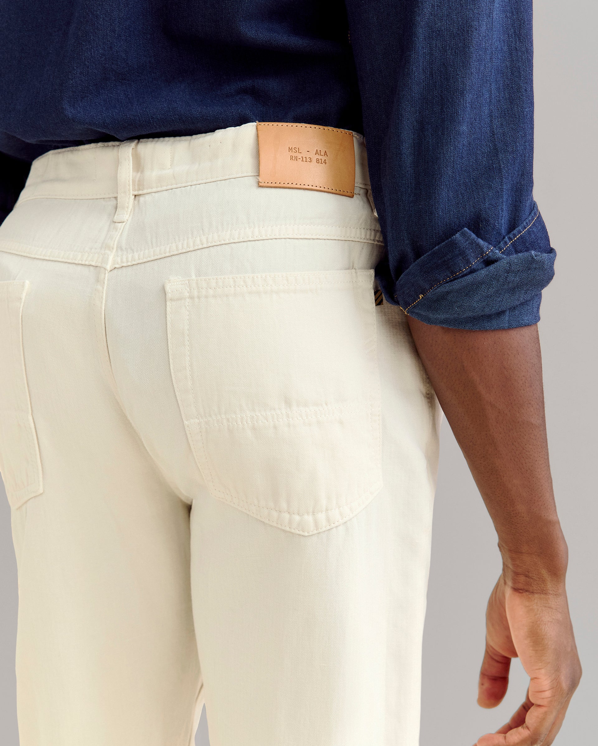 A person wearing a dark blue long-sleeve shirt and off-white jeans is shown from the waist down, focusing on the back pocket and a tan leather patch on the waistband.
