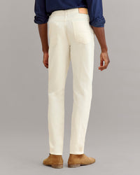 A person stands facing away, wearing a dark blue shirt, light cream jeans, and tan suede shoes, against a plain gray background.
