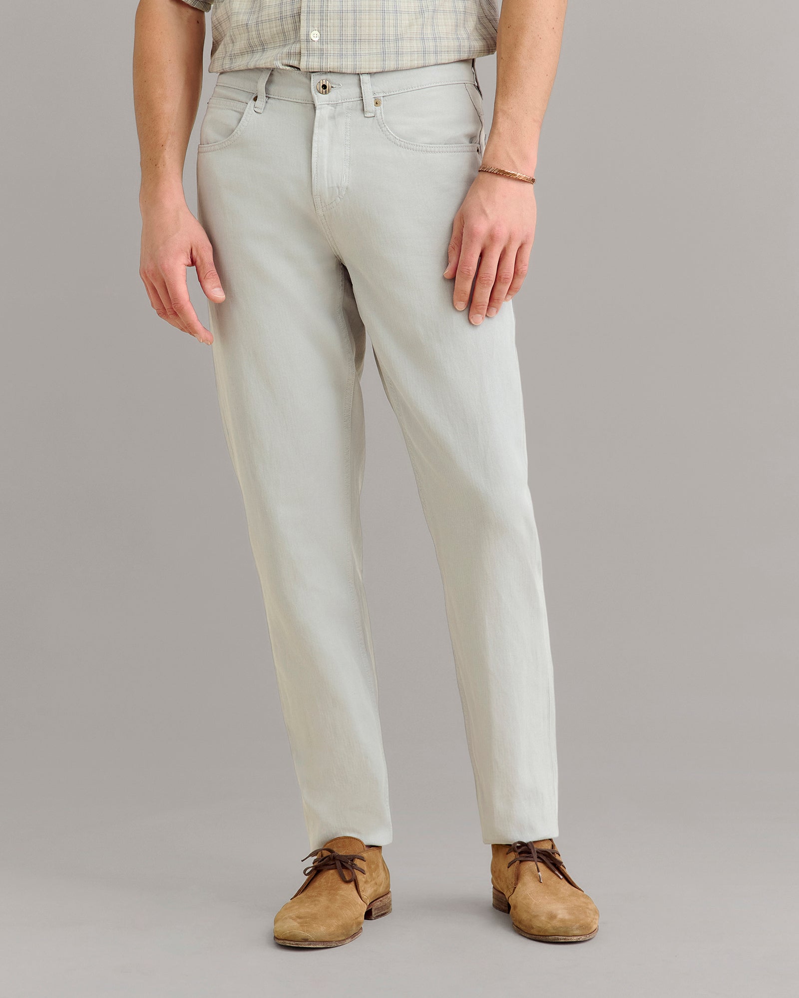 A person wearing light gray jeans, a light plaid shirt, brown suede shoes, and a bracelet, standing against a plain light gray background. Only the body from shoulders down is visible.