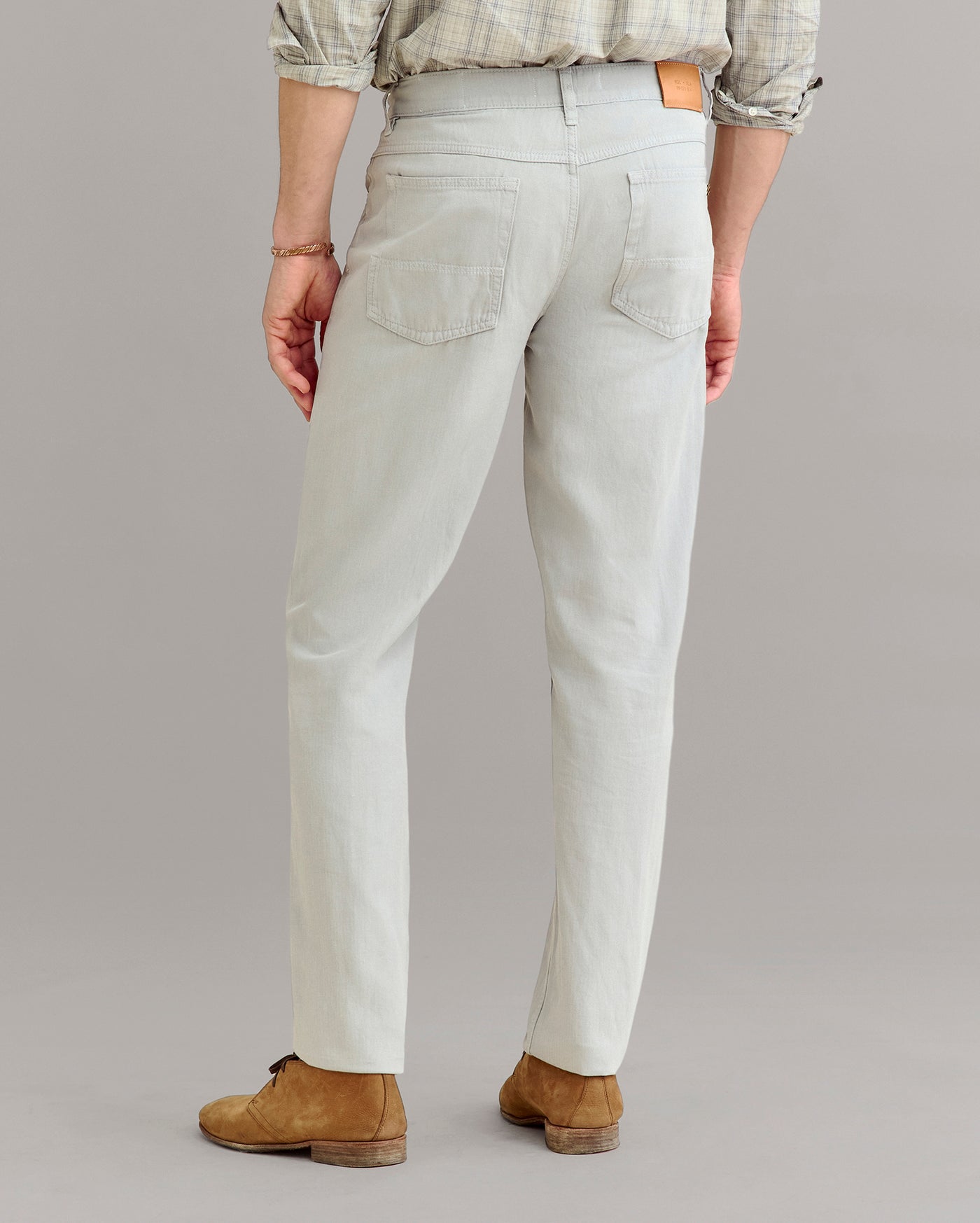 A person wearing light gray jeans, a plaid shirt with rolled sleeves, and brown suede shoes is shown from the back, standing on a plain gray background.