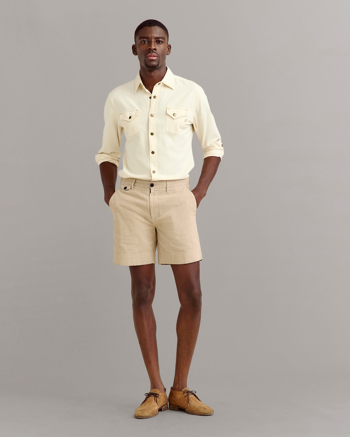 A man stands against a plain gray background wearing a light yellow button-up shirt with rolled-up sleeves, beige shorts, and tan shoes. His hands are in his pockets and he looks forward.