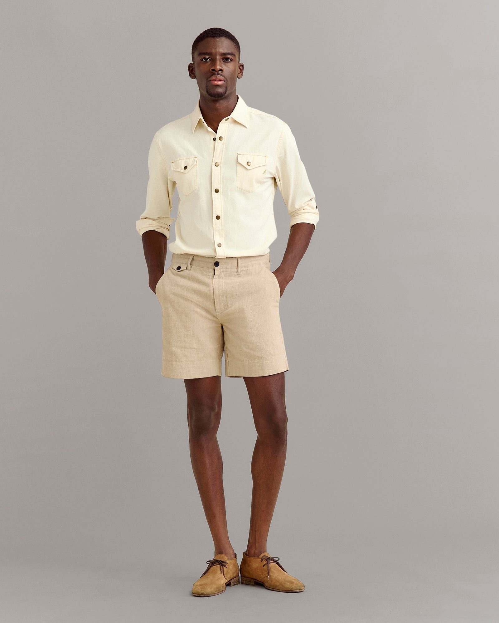 A man stands against a plain gray background wearing a light yellow button-up shirt with rolled-up sleeves, beige shorts, and tan shoes. His hands are in his pockets and he looks forward.