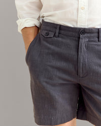 A person wearing a white button-up shirt and textured dark gray shorts stands with one hand in their pocket against a plain background.