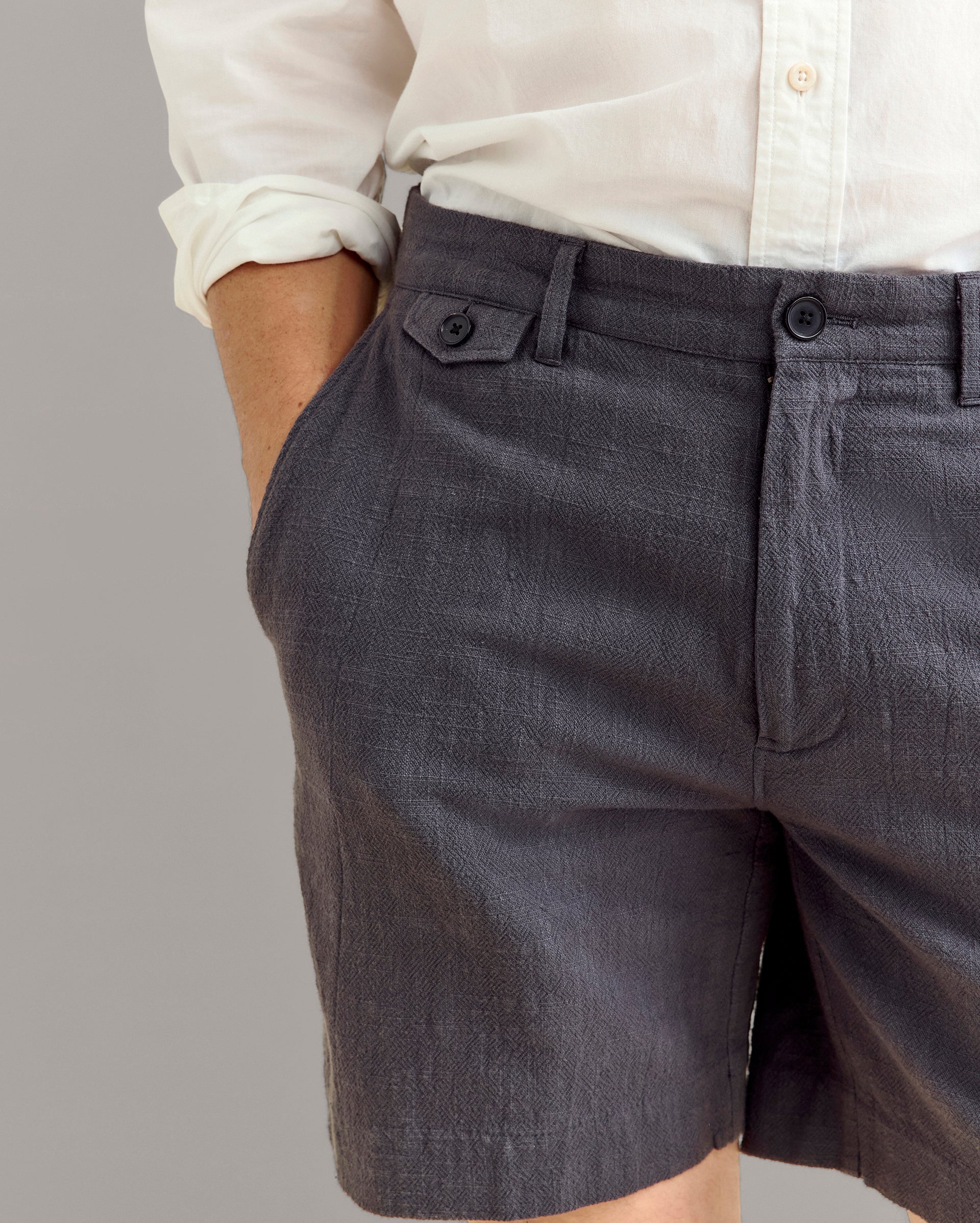 A person wearing a white button-up shirt and textured dark gray shorts stands with one hand in their pocket against a plain background.